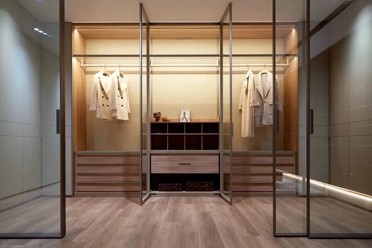 義大利更衣室玻璃門片衣櫃設計，鋁框折疊門開啟後透視木皮吊掛區與抽屜收納，兼具通透展示與開放式衣櫃視覺輕盈感